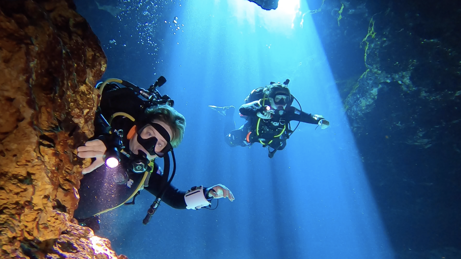Two divers in a cavern with sunlight penetrating the water behind them